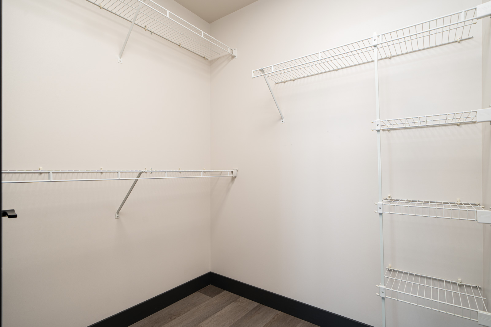 Empty walk-in closet with white wire shelving on two walls, light-colored walls, and wood-style flooring.