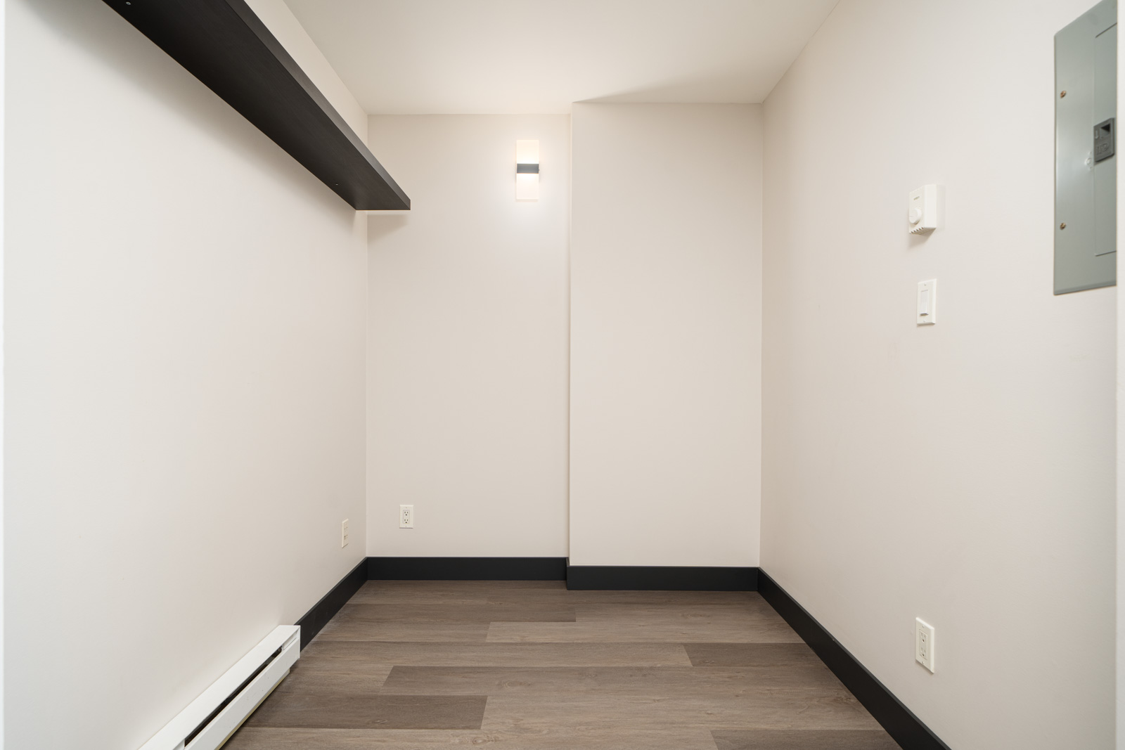 Empty, narrow room with light-colored walls, a dark shelf on the left wall, wood flooring, and a wall-mounted light fixture. There are several electrical outlets and a panel.