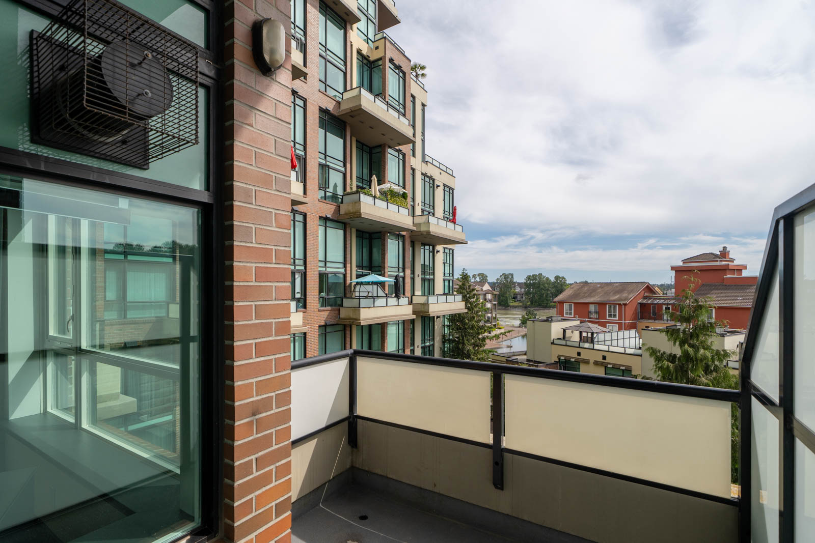 View from a modern apartment balcony overlooking neighboring buildings, with glass railings, brick walls, and a partly cloudy sky.