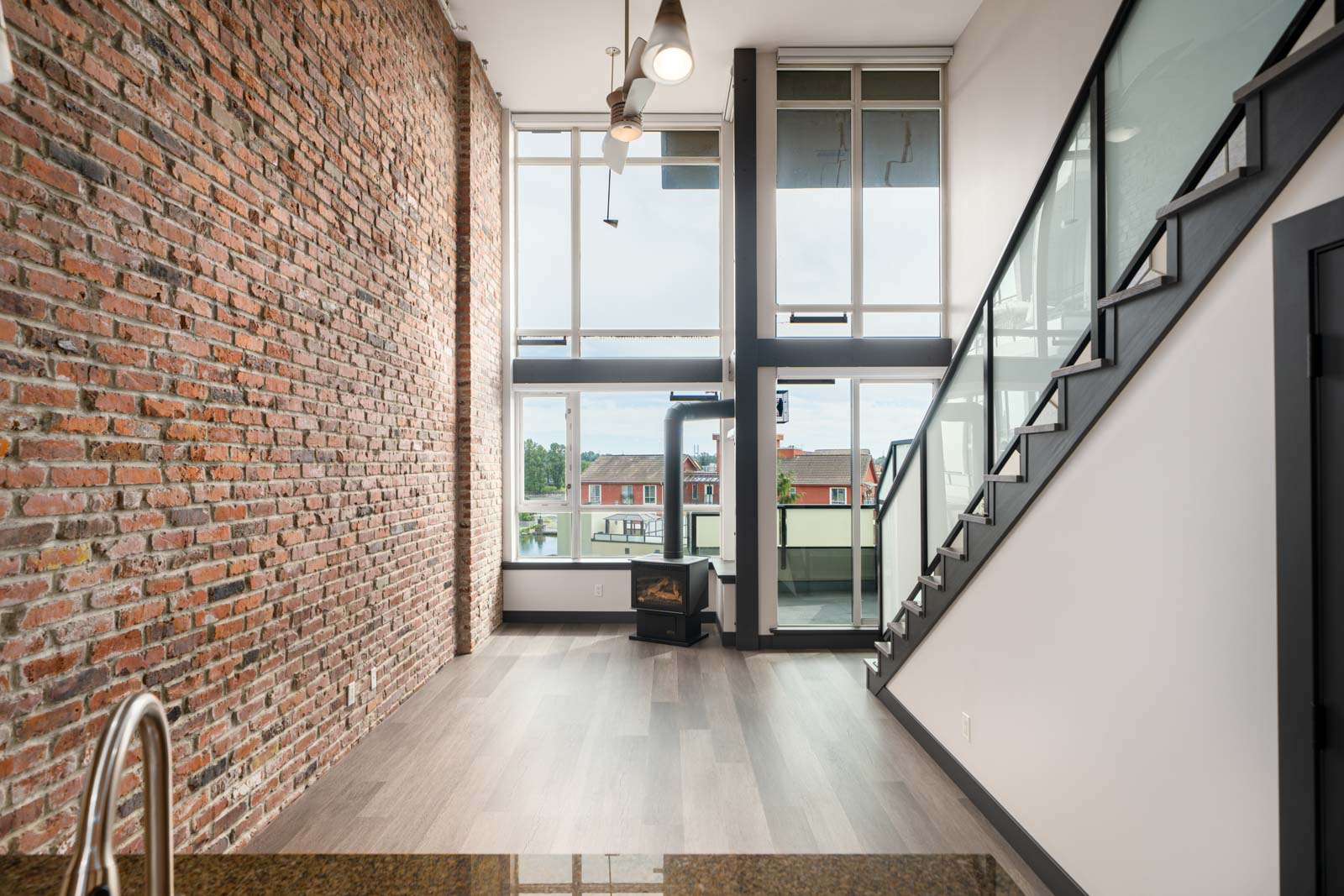Modern loft-style living space with exposed brick wall, large windows, high ceilings, a staircase, and a small fireplace near the windows.