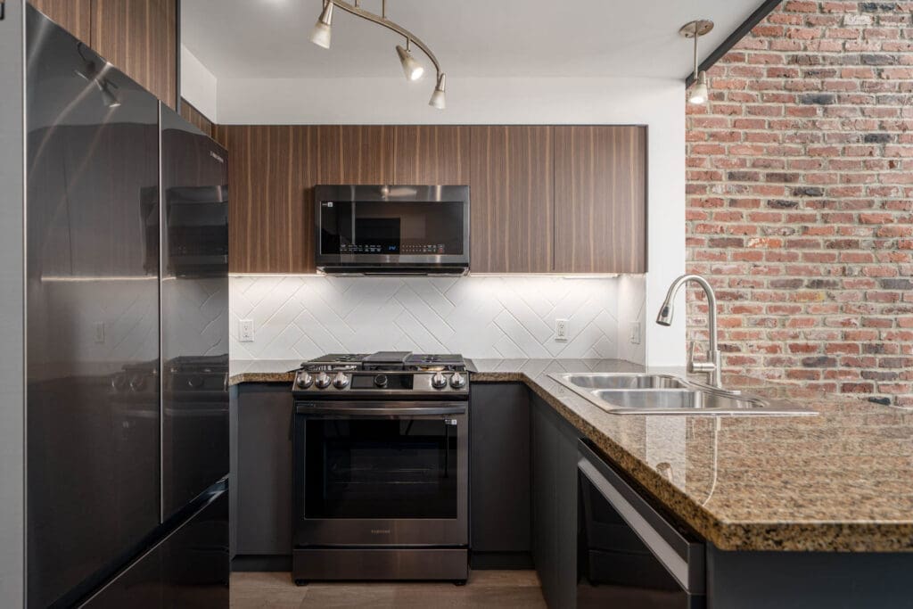 Modern kitchen with stainless steel appliances, brown cabinets, granite countertop, double sink, and an exposed brick wall.