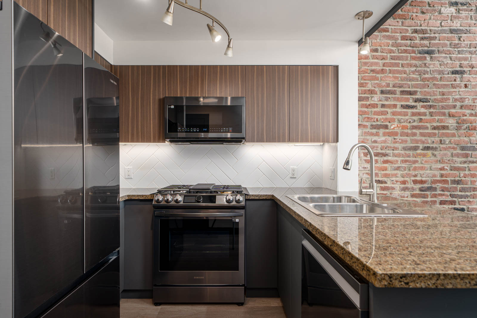 Modern kitchen with stainless steel appliances, brown cabinets, granite countertop, double sink, and an exposed brick wall.