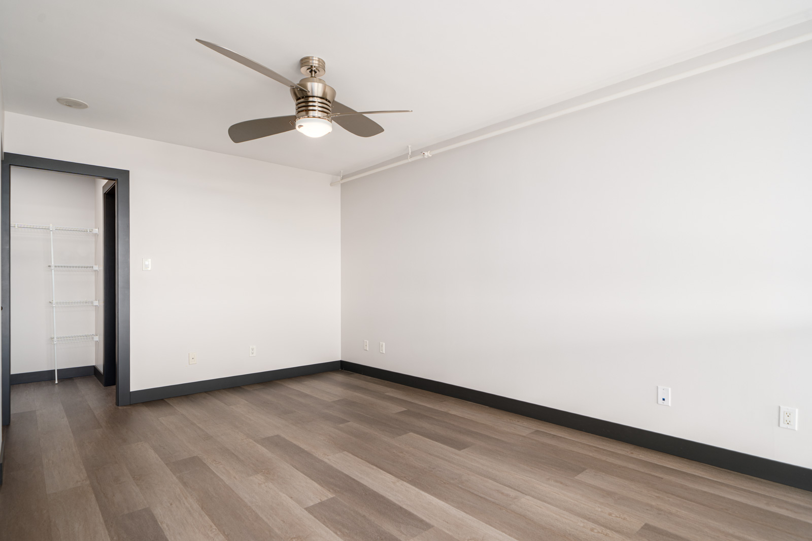 Empty room with light wood flooring, white walls, a silver ceiling fan, and an open doorway leading to a closet with wire shelves.
