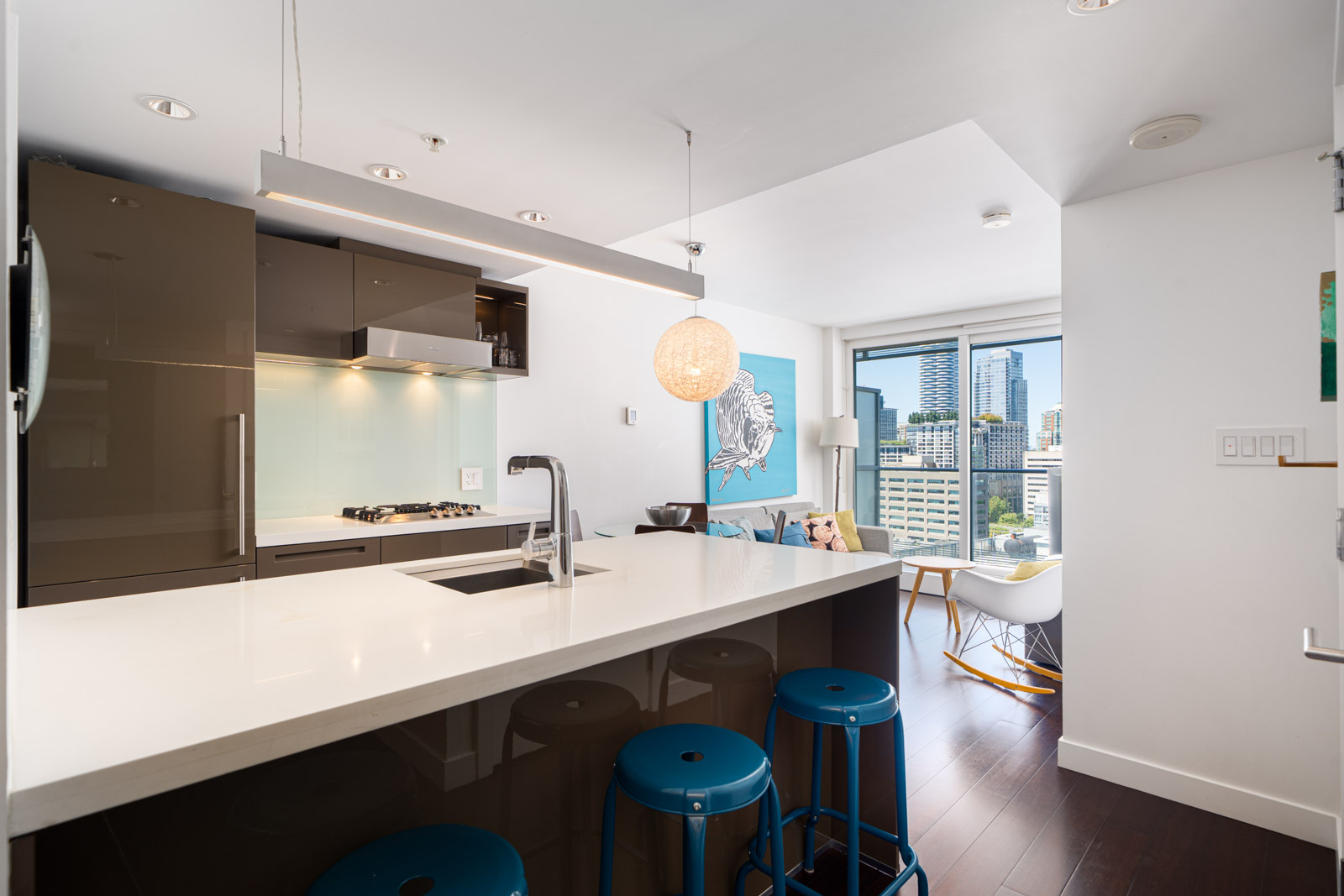 Modern kitchen with white countertops, glossy brown cabinets, and three blue stools, opening to a living area with large windows and cityscape view.