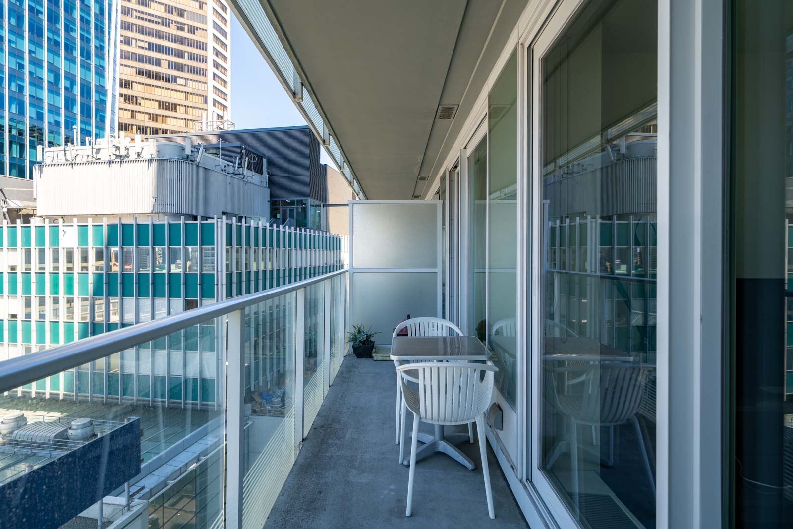 A modern apartment balcony with glass railing, two white chairs, and a small table overlooks nearby office buildings.