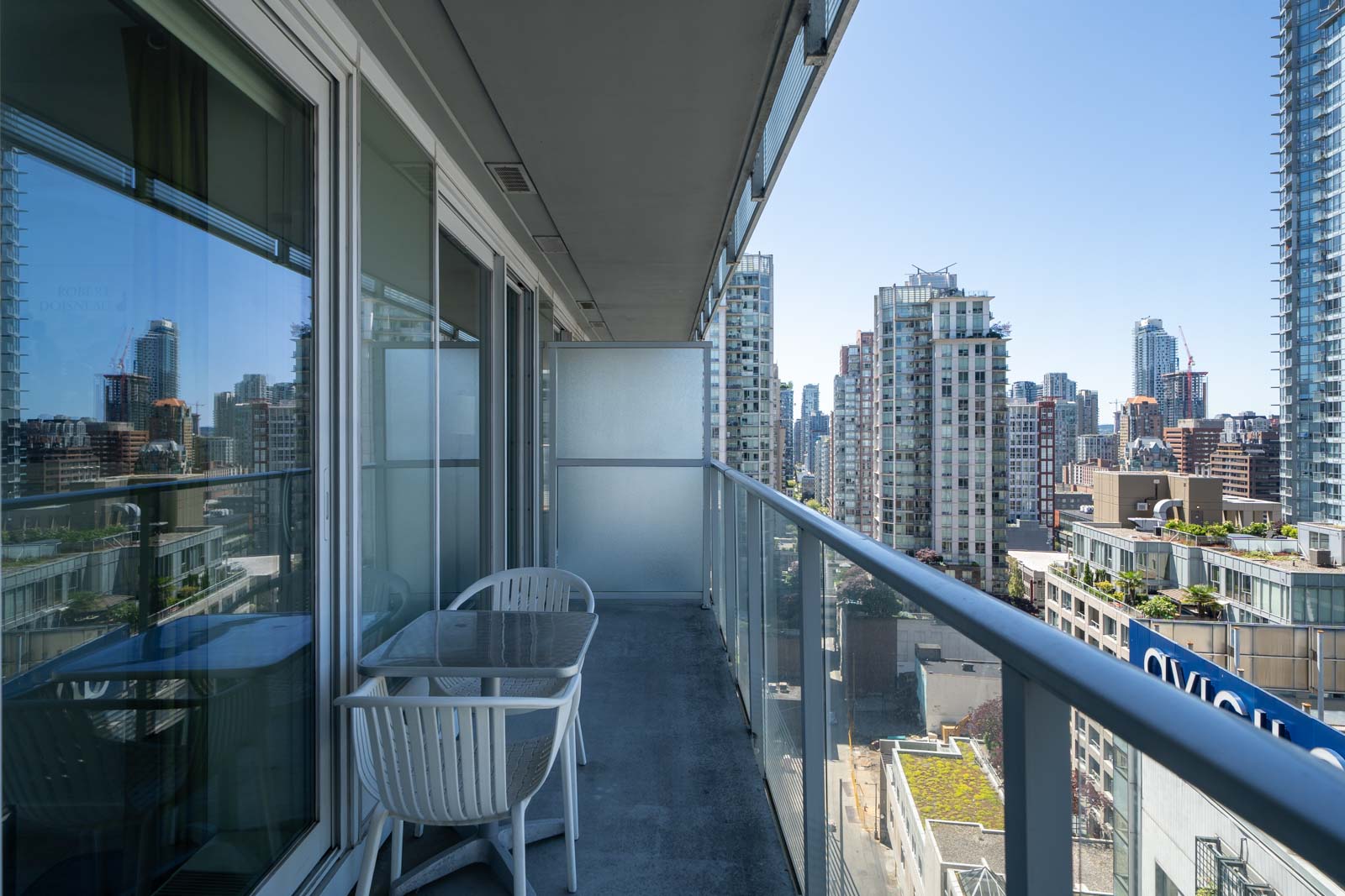 A balcony with a glass railing, a small table, and two chairs overlooks a city skyline with modern high-rise buildings on a clear day.