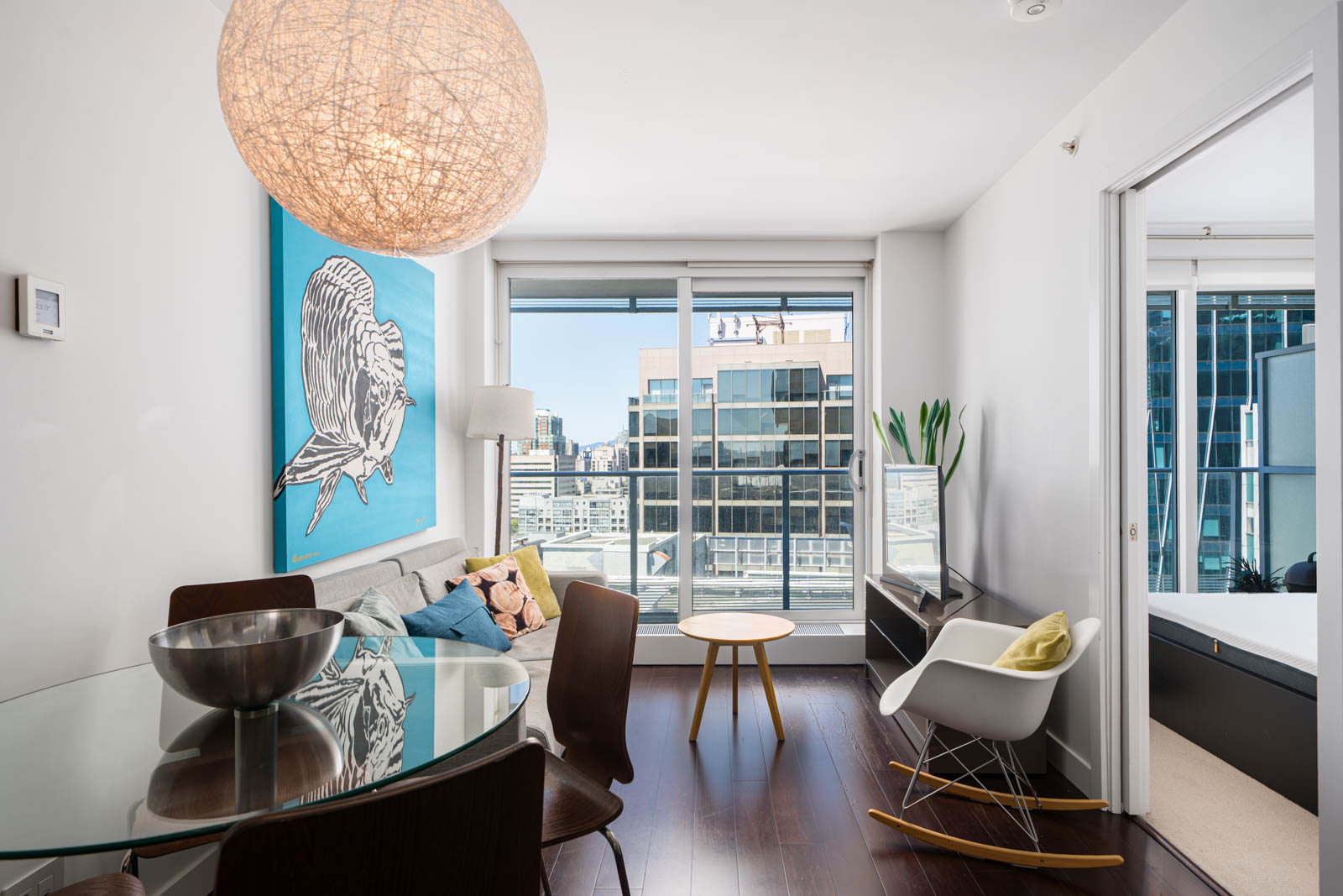 Modern living room with glass dining table, large artwork on the wall, rocking chair, and floor-to-ceiling windows overlooking city buildings. Bedroom visible through open door.