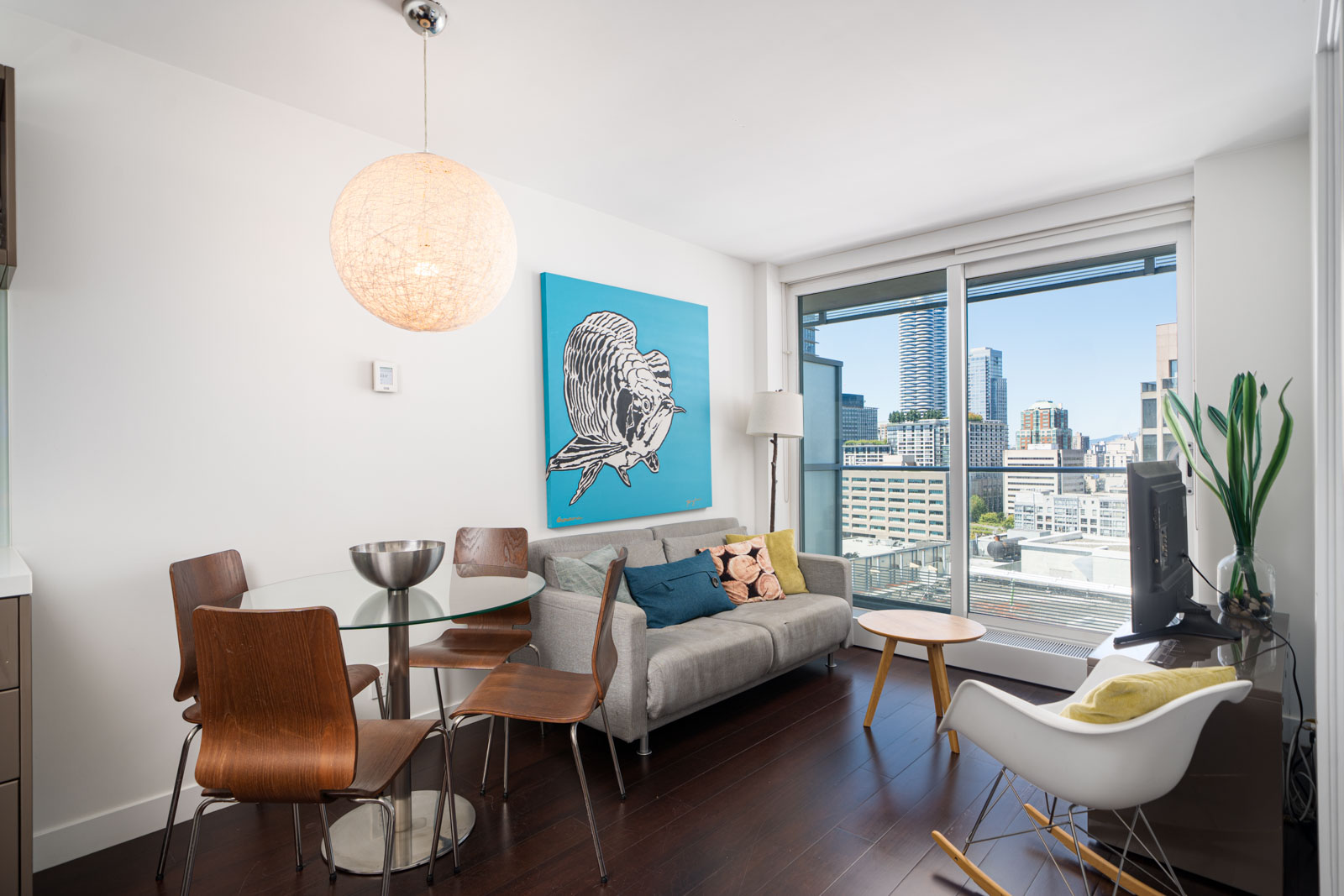 Modern apartment living room with a small dining table, chairs, sofa, wall art, floor lamp, TV, and large window overlooking a city skyline.