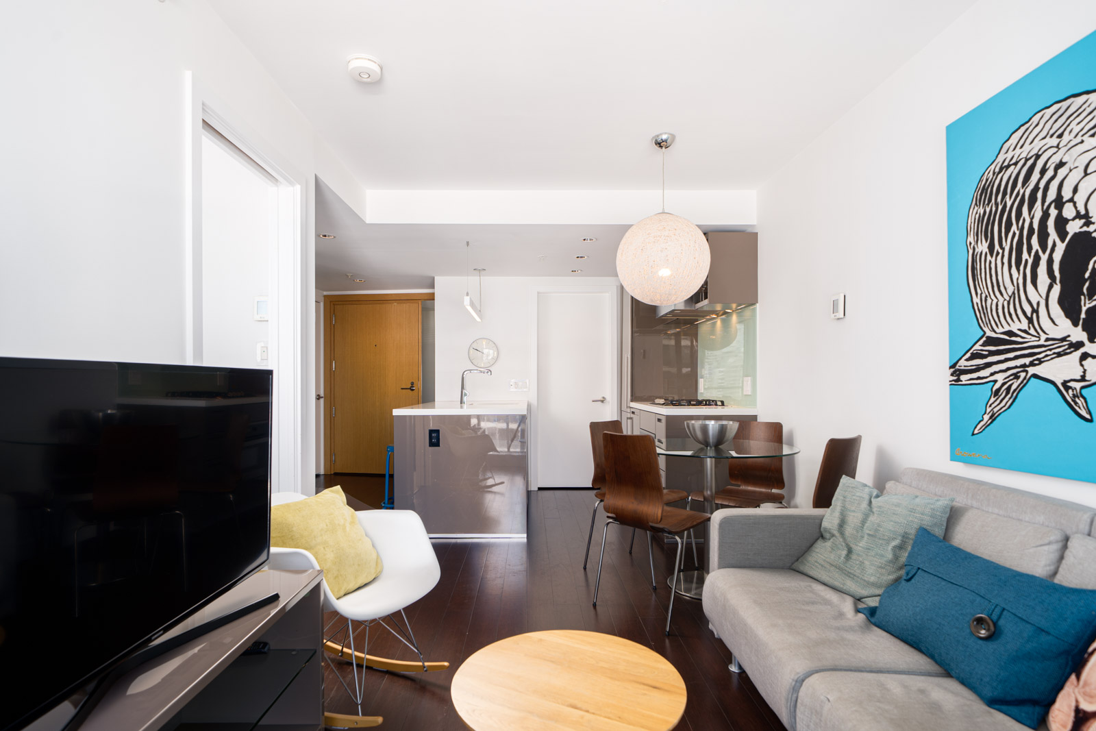 Modern apartment interior featuring a gray sofa, round wooden table, TV, dining area with four chairs, kitchenette, and a large blue artwork on the wall.