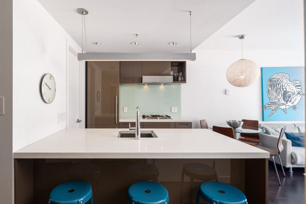 Modern kitchen with a white island, three blue stools, a built-in sink, wall clock, and open view to a dining and living area with contemporary decor.