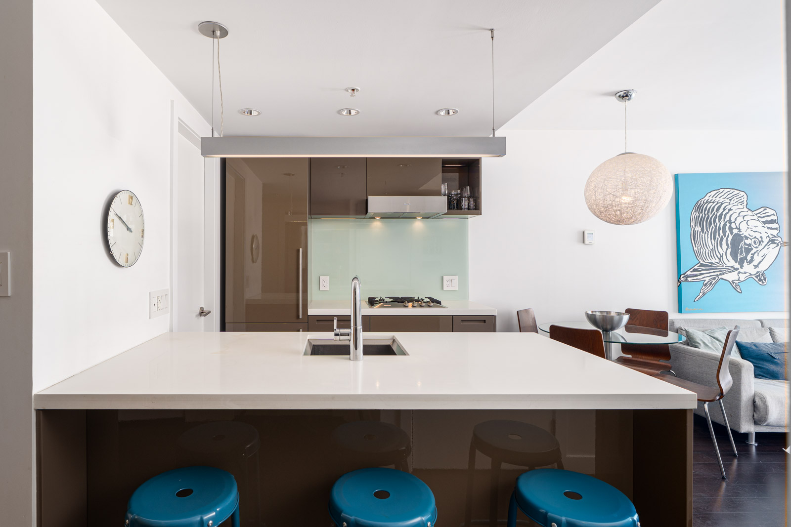 Modern kitchen with a white island, three blue stools, a built-in sink, wall clock, and open view to a dining and living area with contemporary decor.