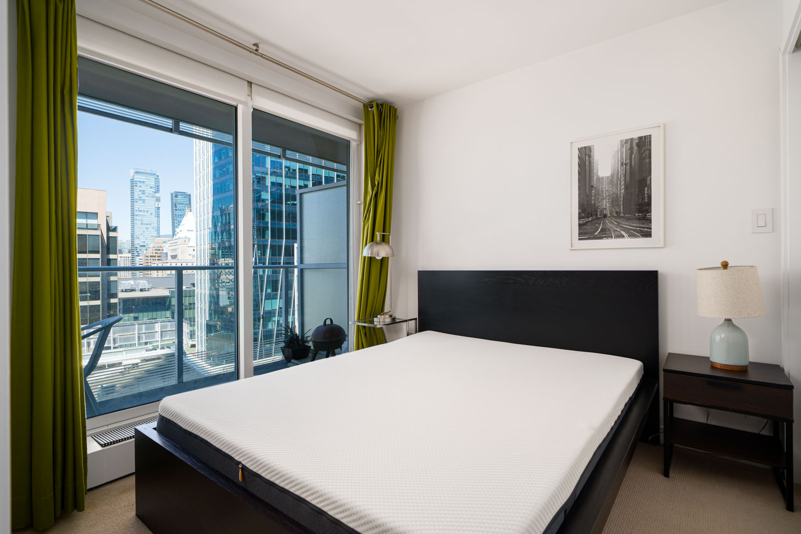 A modern bedroom with a large bed, nightstand with a lamp, framed art on the wall, and floor-to-ceiling window showing cityscape and buildings outside.