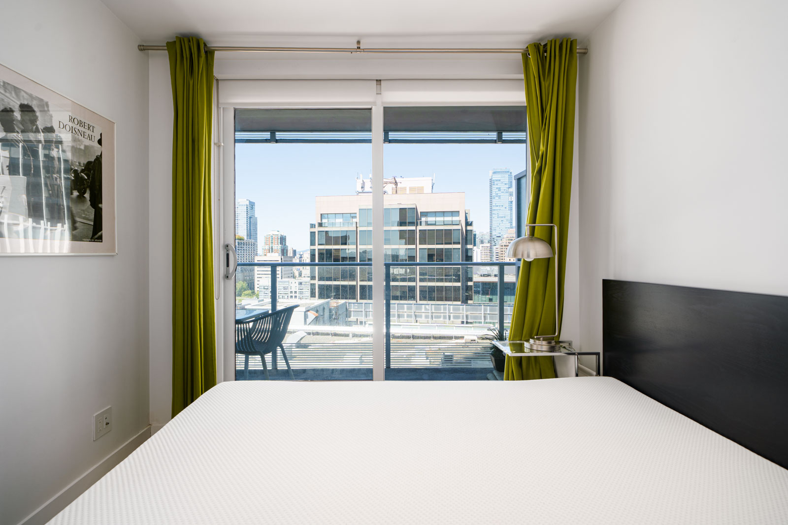A modern bedroom with a large window, green curtains, a bed with a white mattress, a side table, and a cityscape view outside.