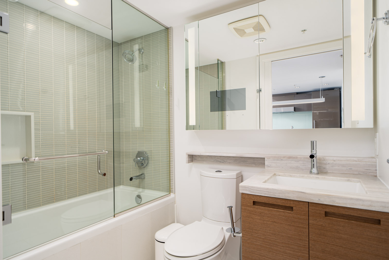 Modern bathroom with glass-enclosed shower and bathtub, wall-mounted toilet, white sink, wood vanity, and mirrored cabinet.
