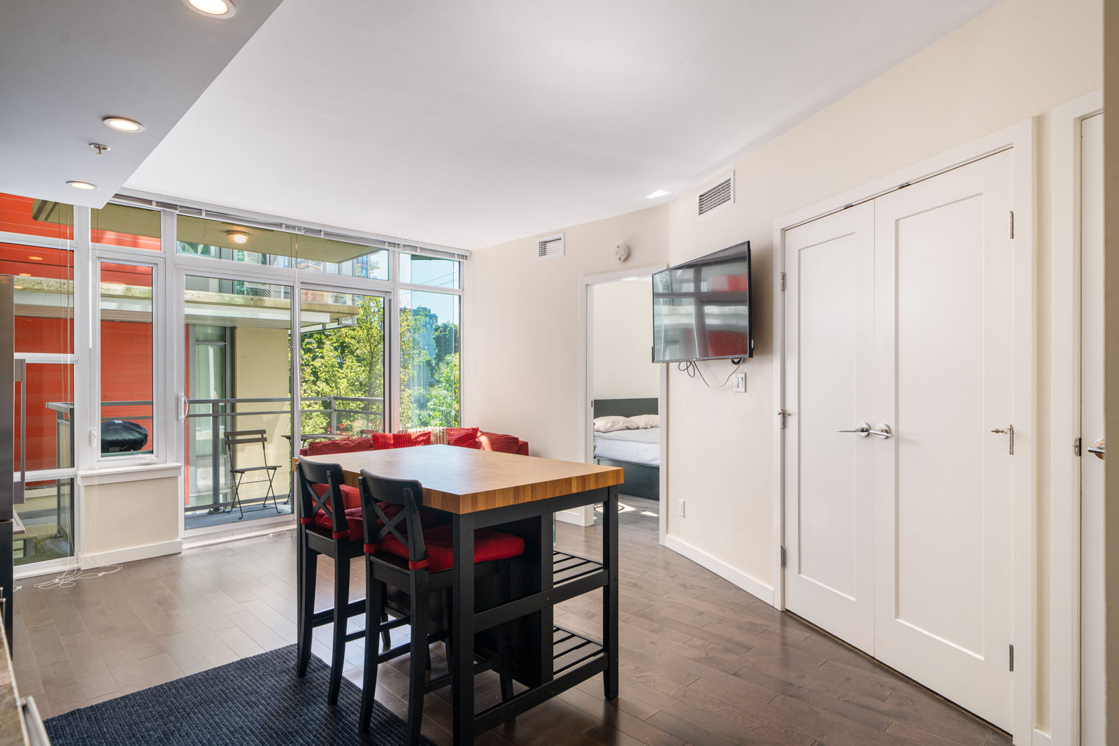 Modern apartment with a dining table and chairs, wall-mounted TV, and balcony access. A bedroom is visible through an open doorway. White walls and large windows provide natural light.