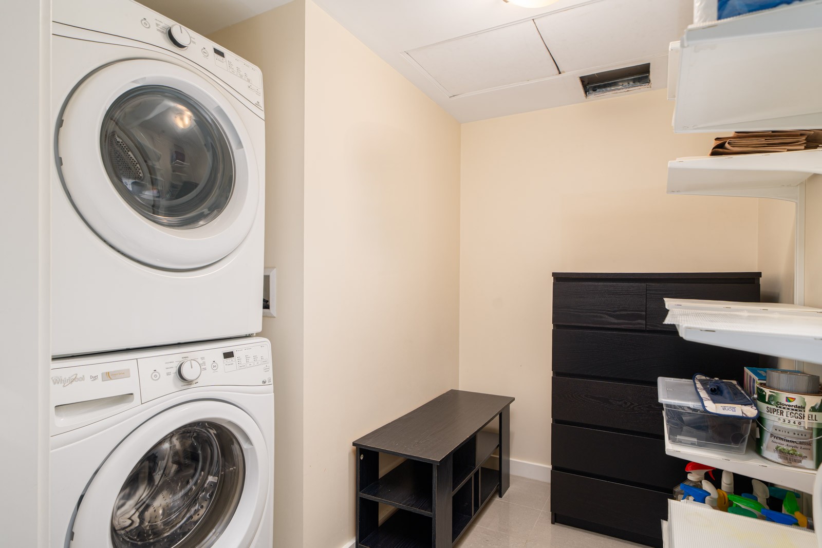 A laundry room with a stacked washer and dryer on the left, black storage furniture, shelves with bins on the right, and beige walls.