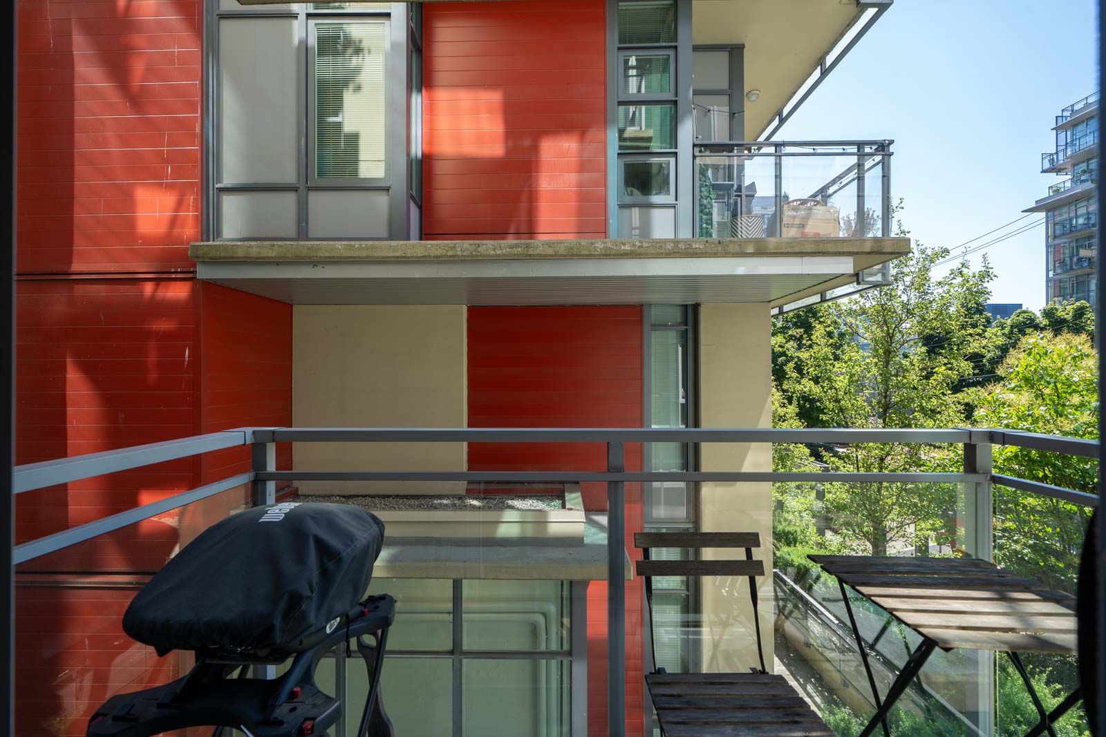 A small balcony with a grill and folding table overlooks modern red and beige apartment buildings, with trees and other buildings in the background.