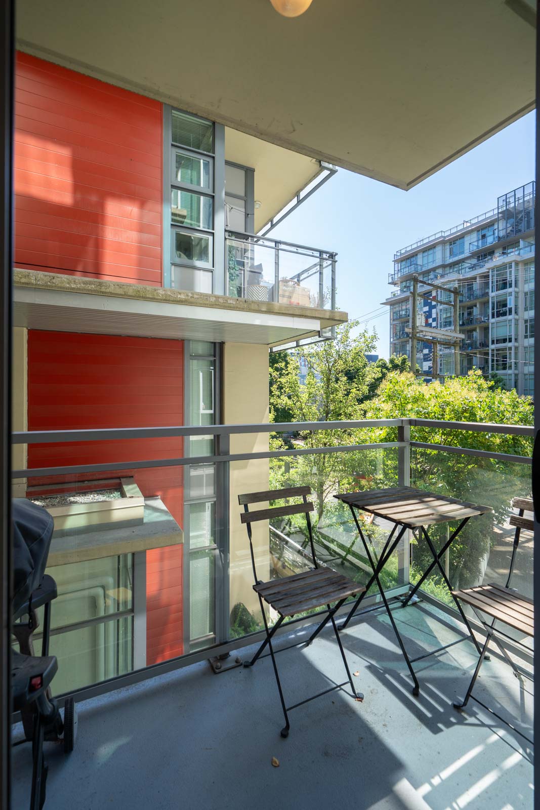 A small balcony with a black metal table and two chairs overlooks neighboring modern apartment buildings and green trees on a sunny day.