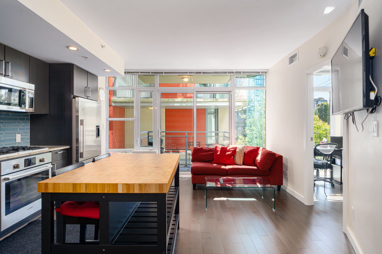 Modern apartment interior with kitchen featuring stainless steel appliances, wooden island, and open living area with a red sofa and large windows leading to a balcony.