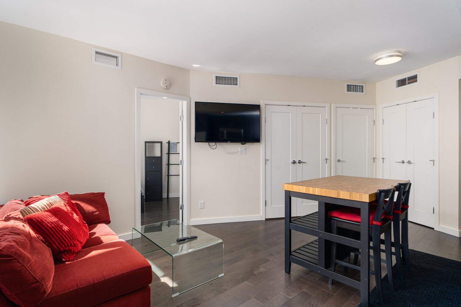 Modern living room with a red sofa, glass coffee table, wall-mounted TV, and a kitchen island with two chairs. Three white doors are visible in the background.