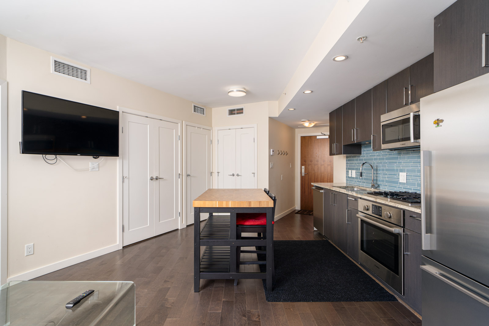 Modern kitchen with stainless steel appliances, dark cabinets, center island, wall-mounted TV, and double doors; open layout with hardwood flooring.