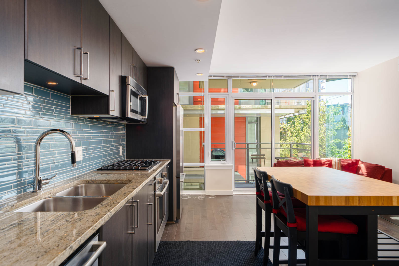 Modern kitchen with dark cabinets, stainless steel appliances, granite countertop, blue tile backsplash, and a dining table with red chairs near large windows.