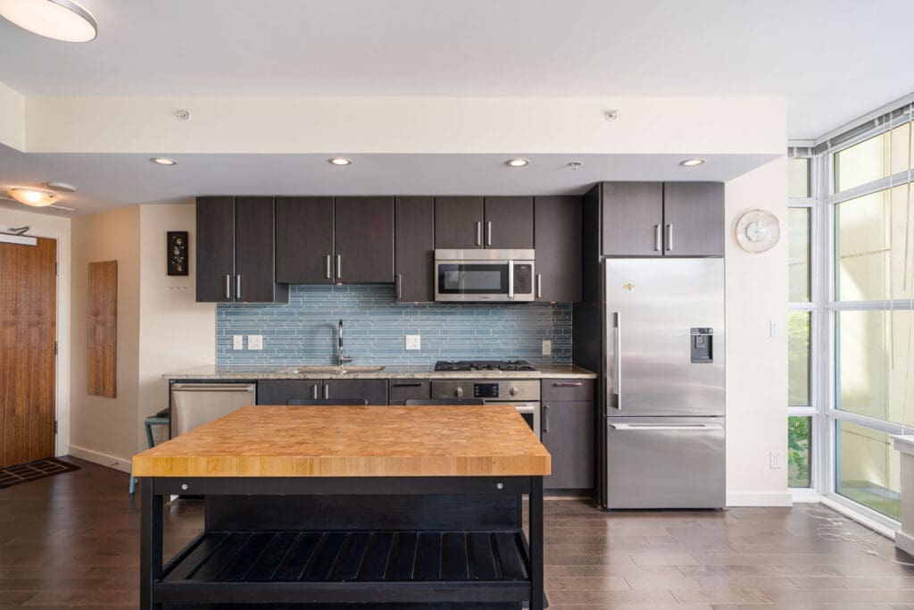 Modern kitchen with dark cabinets, stainless steel appliances, blue tile backsplash, a wooden island, and large windows letting in natural light.