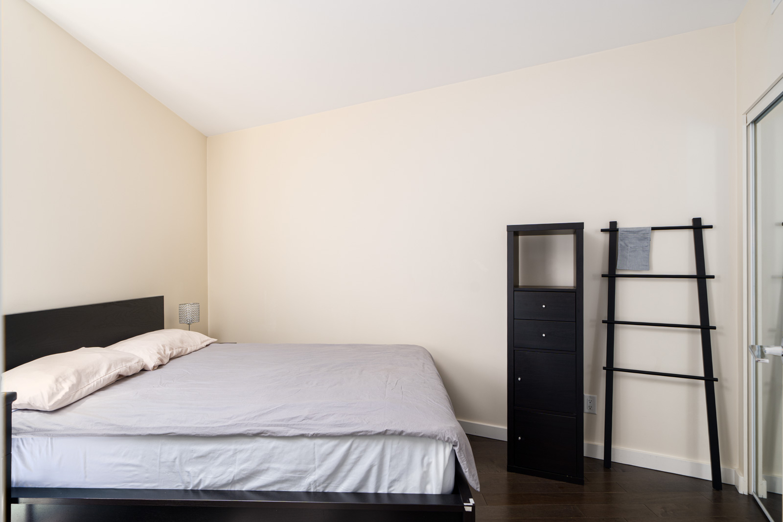 Minimalist bedroom with a neatly made bed, two pillows, a black dresser, and a decorative black ladder against cream-colored walls.