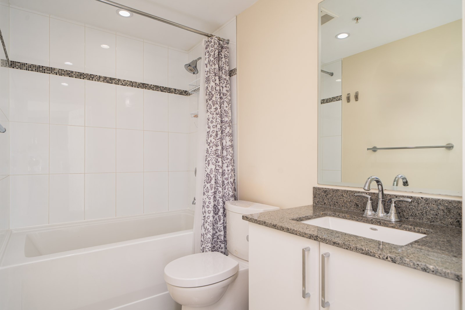 Modern bathroom with a bathtub and shower, white tiles, a patterned shower curtain, toilet, and a vanity with a granite countertop and mirror.