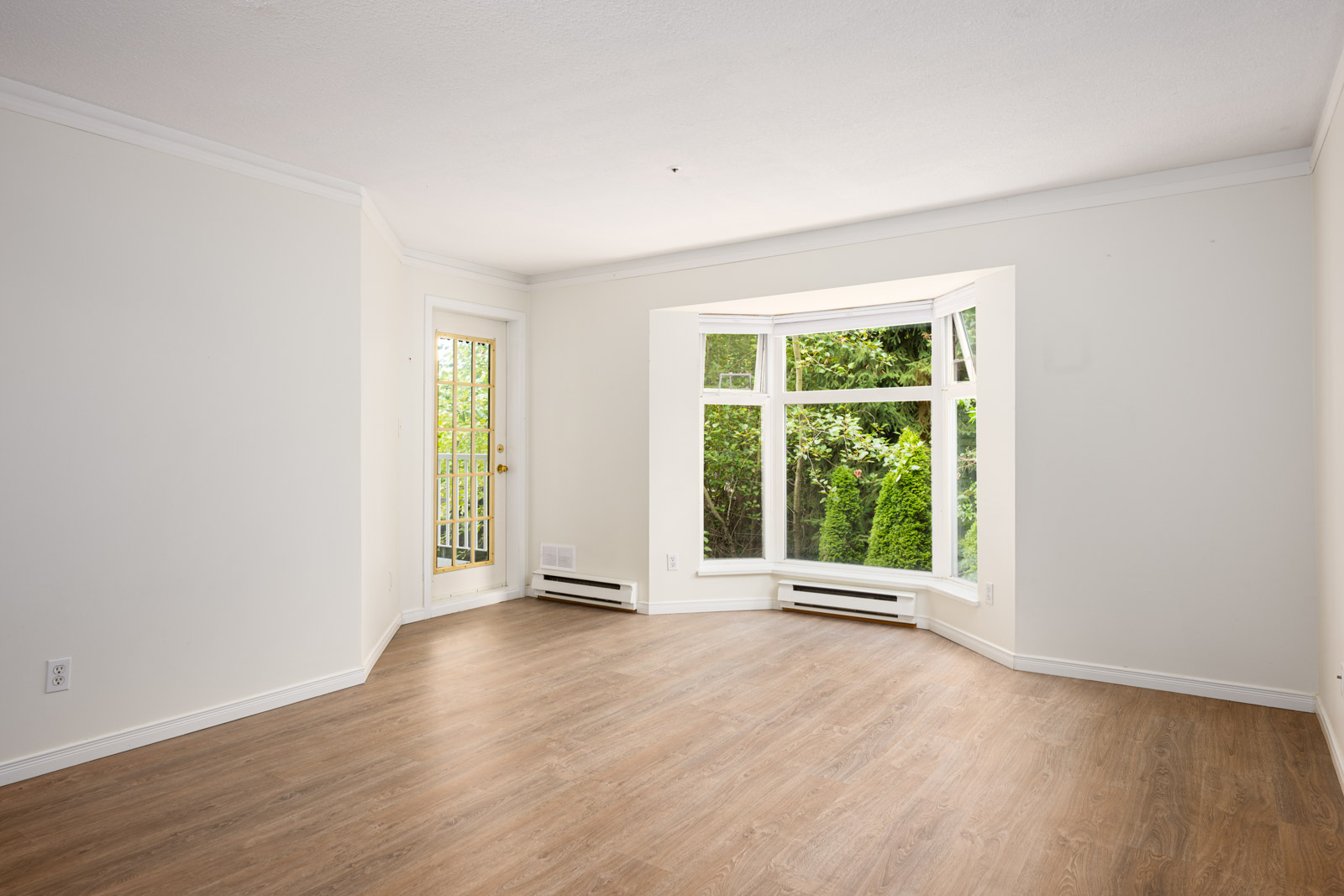 Empty room with white walls, wood flooring, a glass door, and a large bay window overlooking greenery outside.