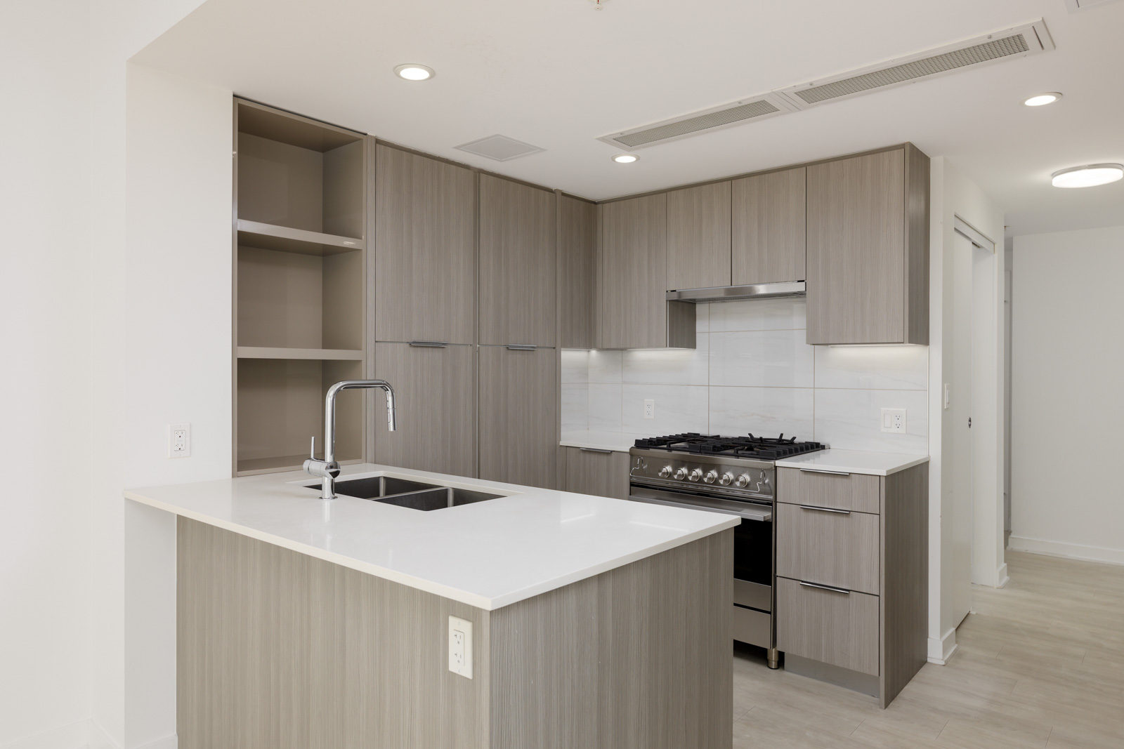 Modern kitchen with light wood cabinets, white countertops, stainless steel appliances, and a central island with a sink under recessed lighting.