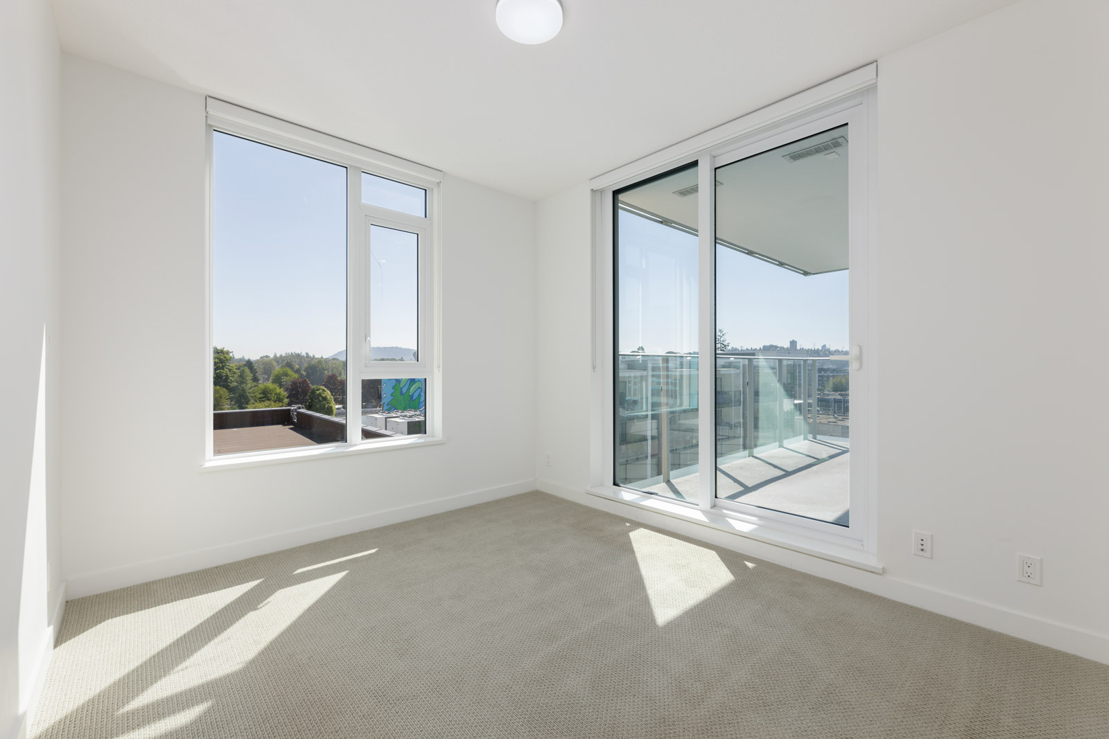 Empty, sunlit room with beige carpet, white walls, a large window, and floor-to-ceiling glass doors leading to a balcony with a view outside.