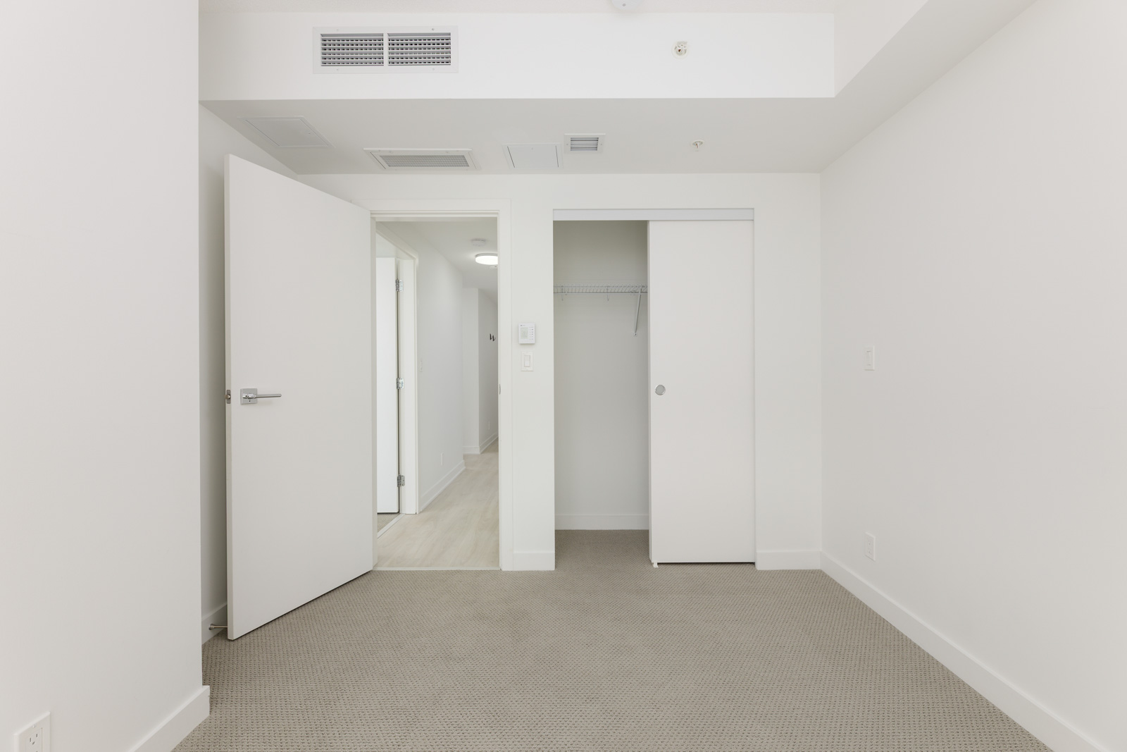 Empty white-walled room with beige carpet, open door leading to hallway, and closet with sliding doors and a single shelf.