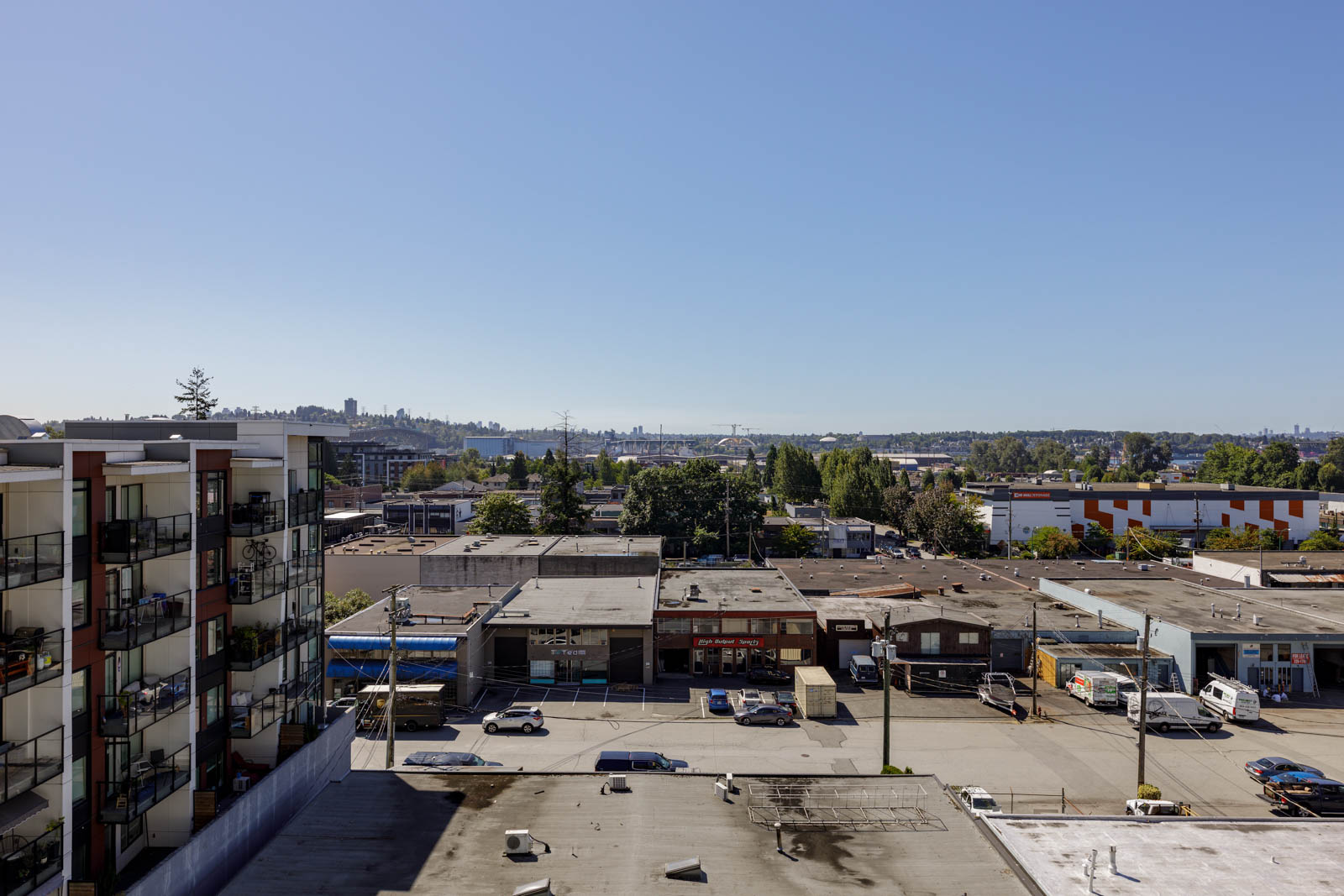 View of a cityscape with apartment buildings on the left, commercial warehouses below, and a distant skyline under a clear blue sky.