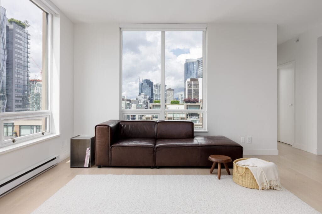 A modern living room with a brown leather sofa, small wooden stool, basket with a blanket, large windows, and a city skyline view.