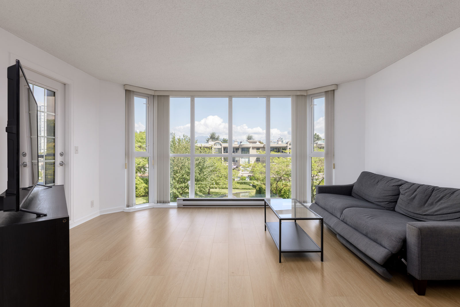 A minimalist living room with a gray sofa, glass coffee table, TV on a stand, light wood flooring, and large windows overlooking greenery and residential buildings.