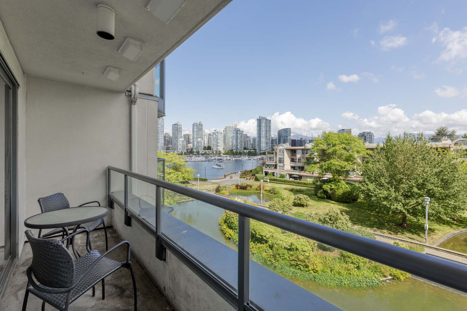 A balcony with a small table and two chairs overlooks a park, marina, and city skyline under a partly cloudy sky.