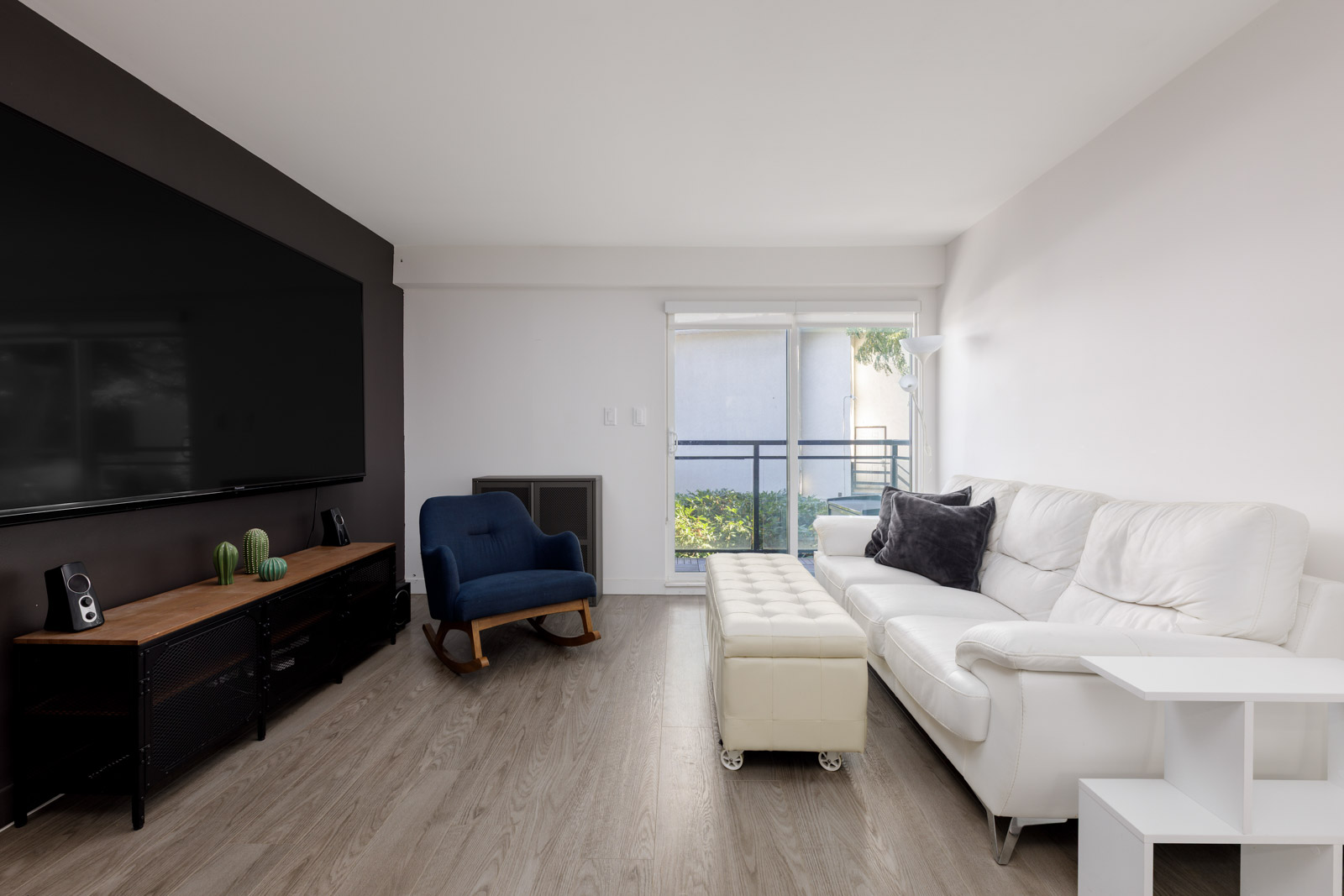Modern living room with a large TV, white sectional sofa, blue armchair, ottoman, wooden TV stand, and sliding glass doors leading to a balcony.