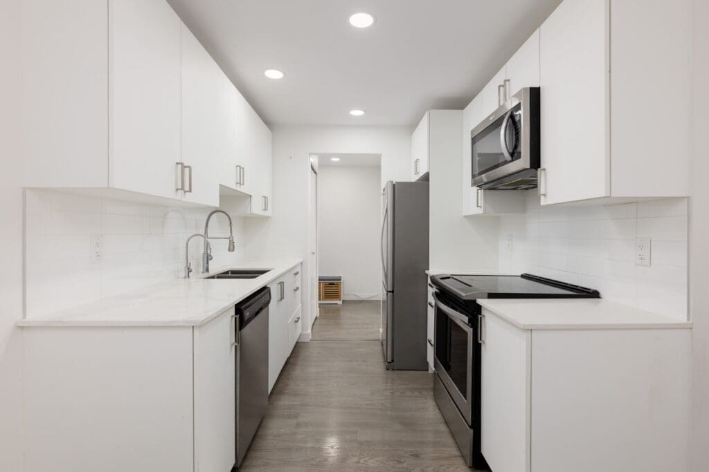 Modern galley kitchen with white cabinets, stainless steel appliances, electric stove, built-in microwave, double sink, and light wood flooring under recessed ceiling lights.