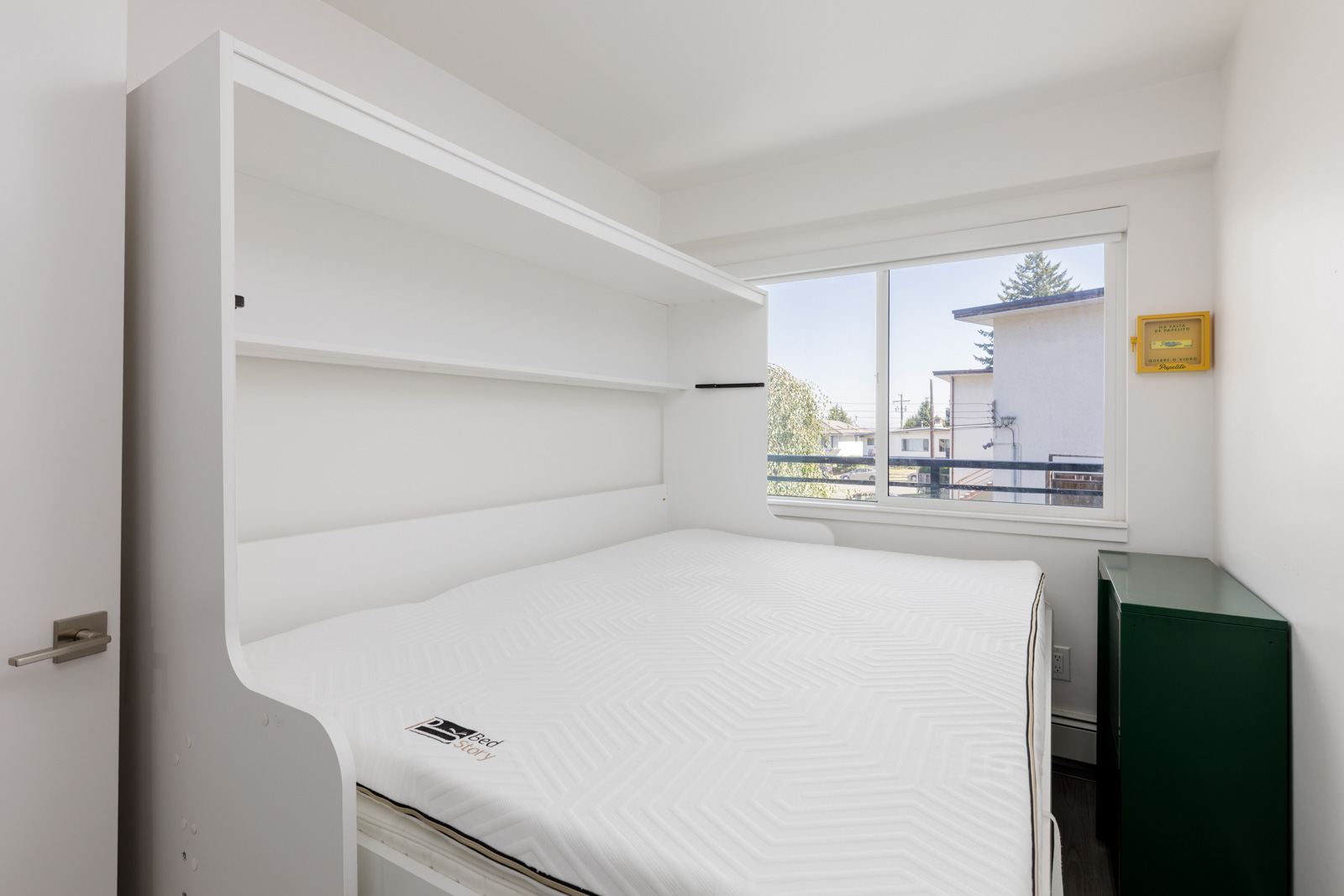 A small, modern bedroom with a Murphy bed, white walls, a large window, and a green cabinet near the corner.