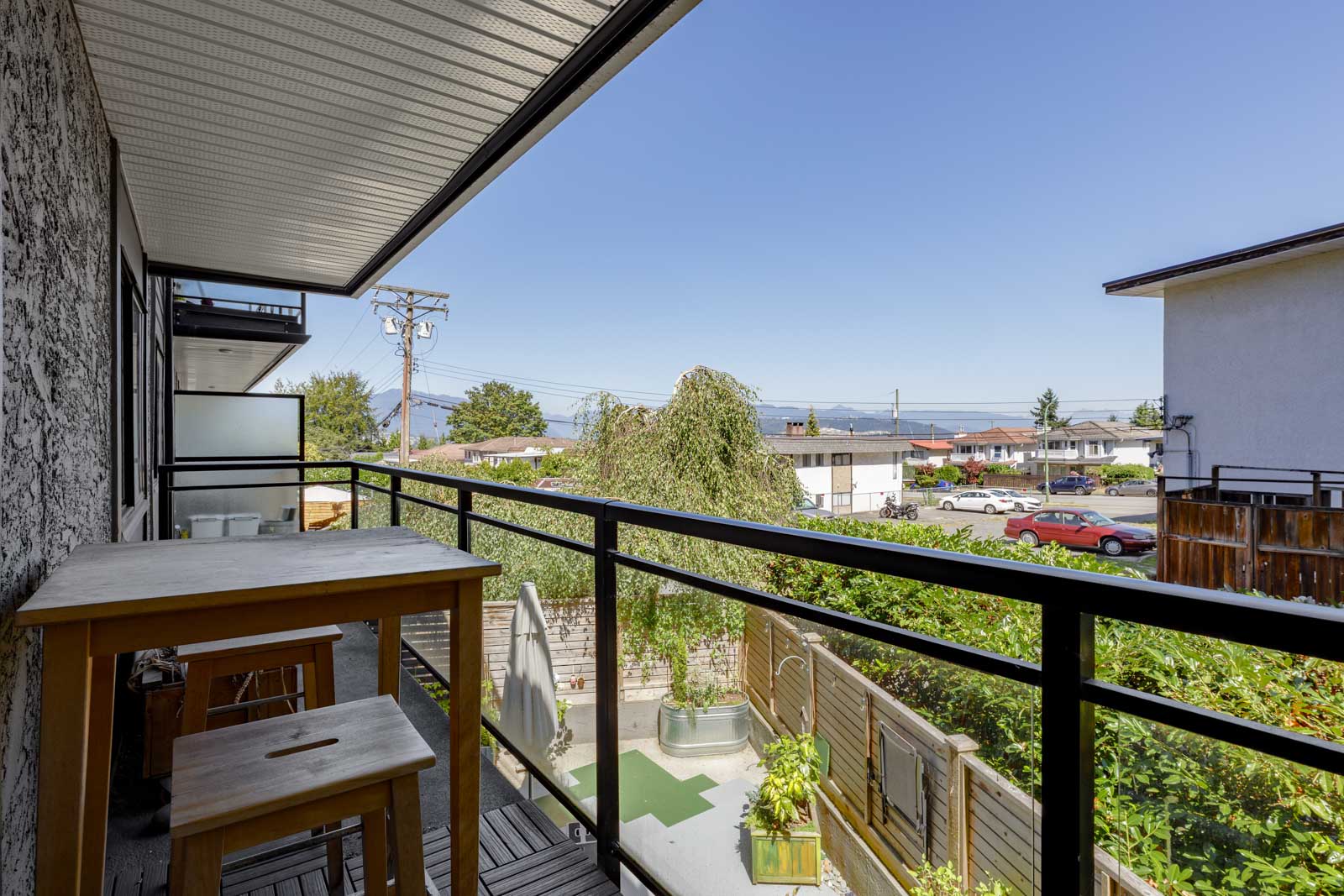 A balcony with a wooden table and stool overlooks a residential neighborhood, with clear skies and distant mountains visible in the background.