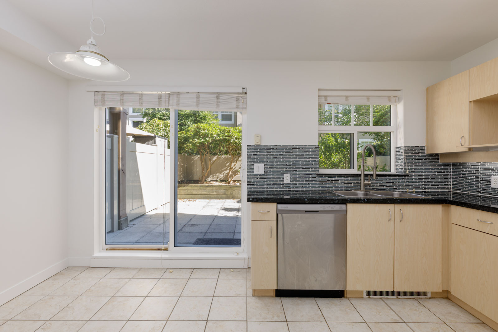 Modern kitchen with light wood cabinets, stainless steel dishwasher, tile floor, and a sliding glass door leading to a patio with a fenced yard.
