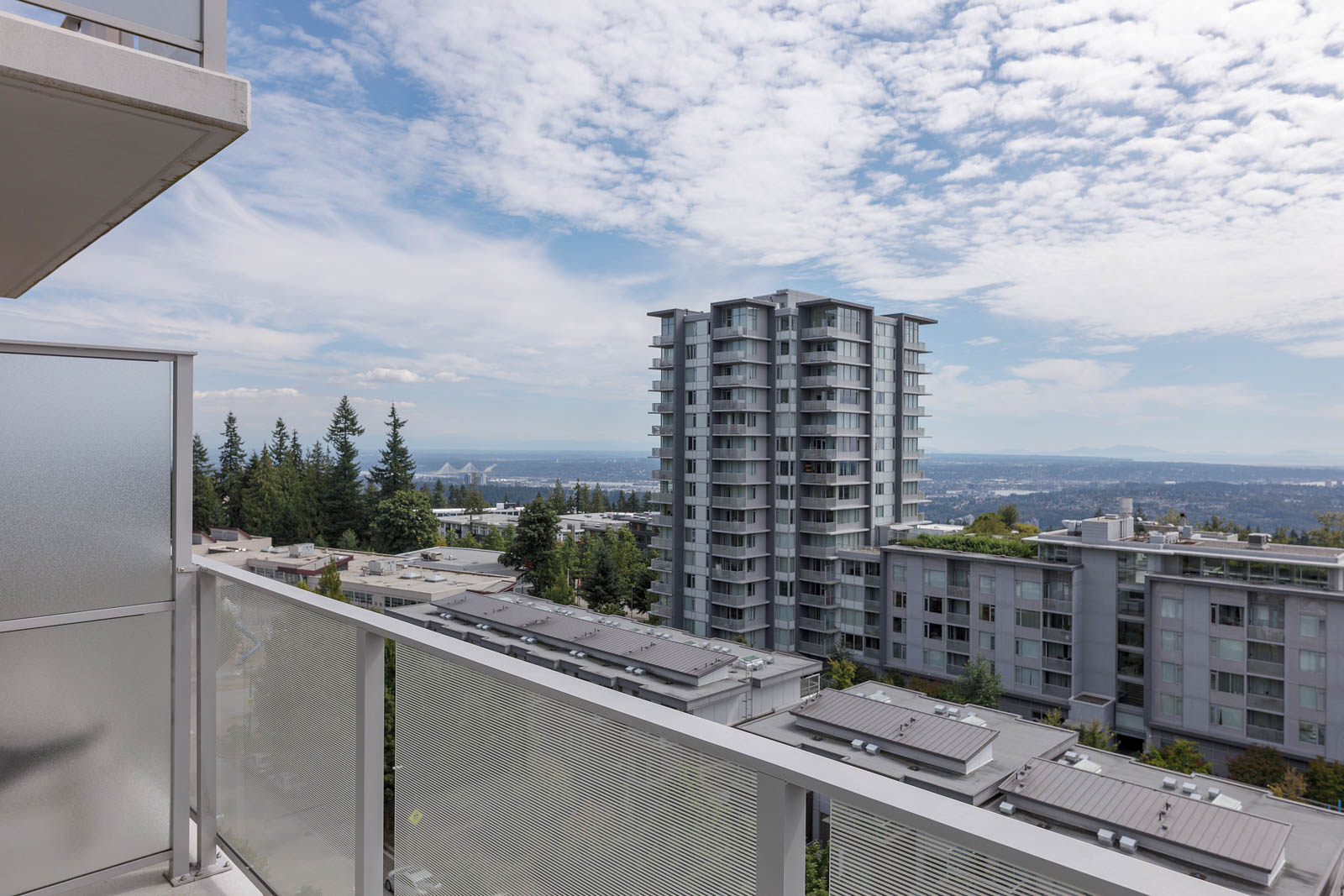 View from a balcony overlooking several modern apartment buildings, trees, and distant hills under a partly cloudy sky.