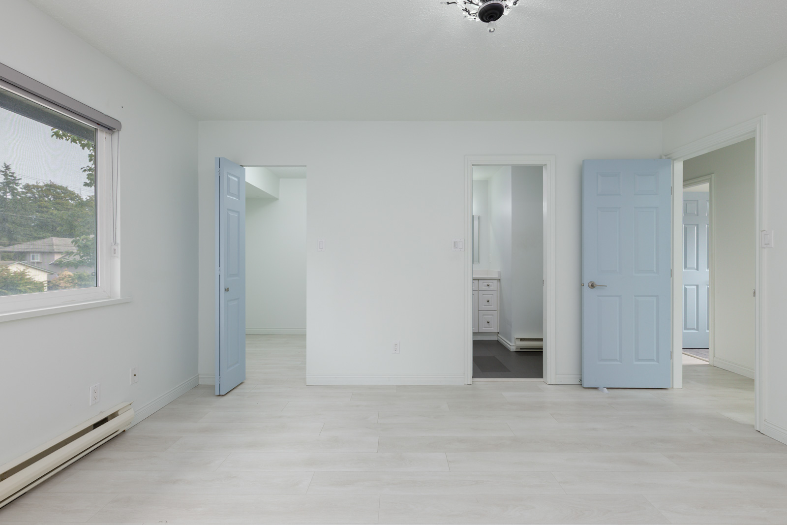 Empty room with light-colored floors, white walls, a window on the left, and three light blue doors leading to adjacent rooms or spaces.