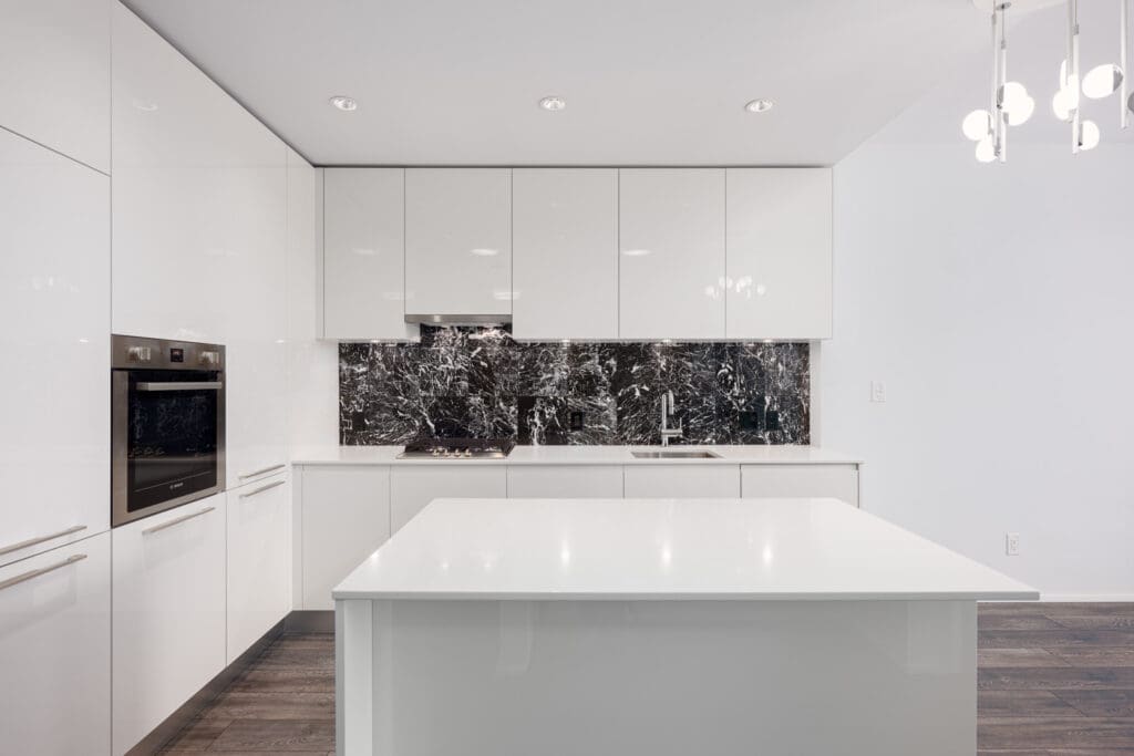 Modern kitchen with glossy white cabinets, marble backsplash, built-in oven, stovetop, sink, and a white island under recessed and pendant lighting.