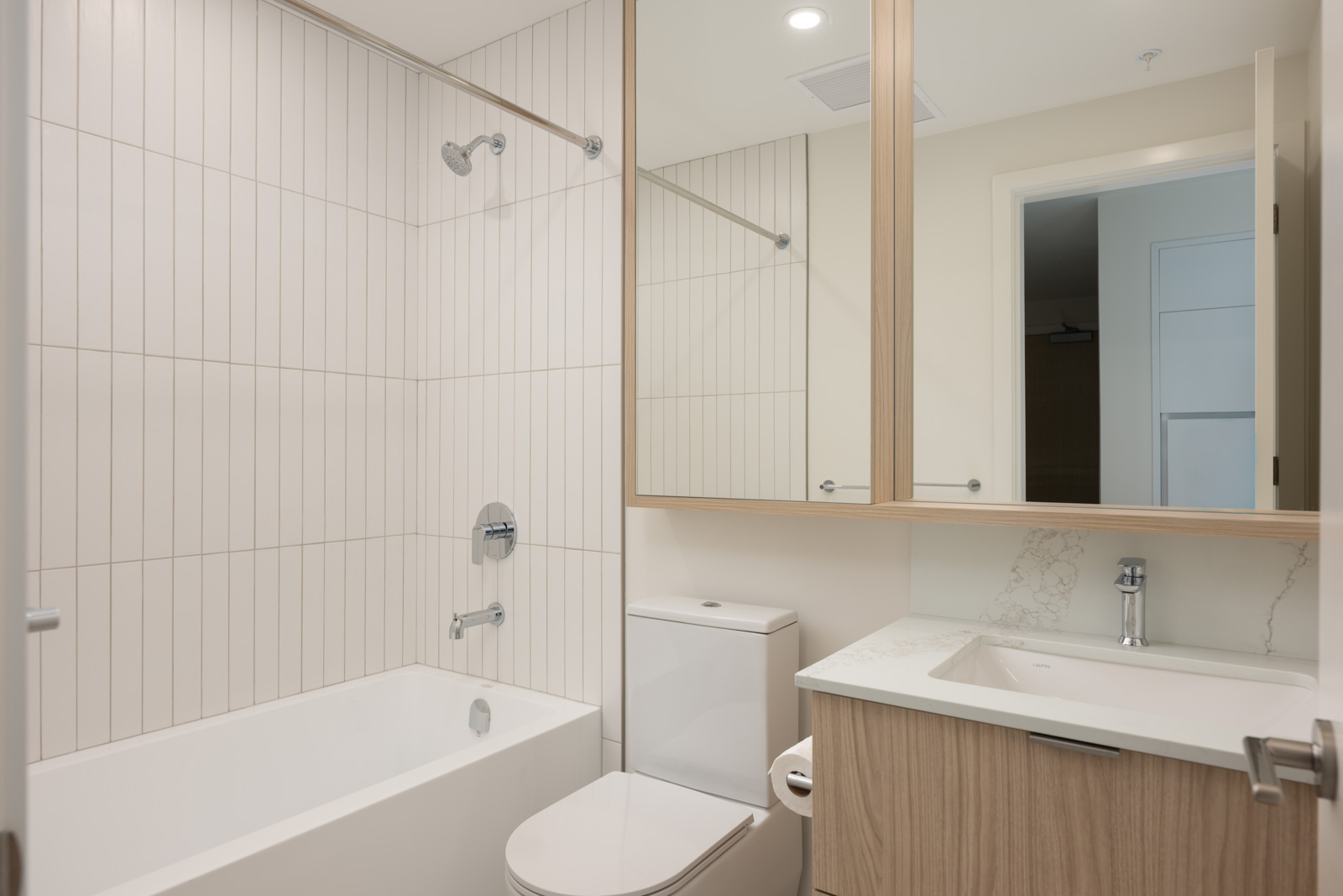 Modern bathroom with a bathtub-shower combo, white tile walls, a toilet, a wood vanity with a white countertop, and a large mirror above the sink.