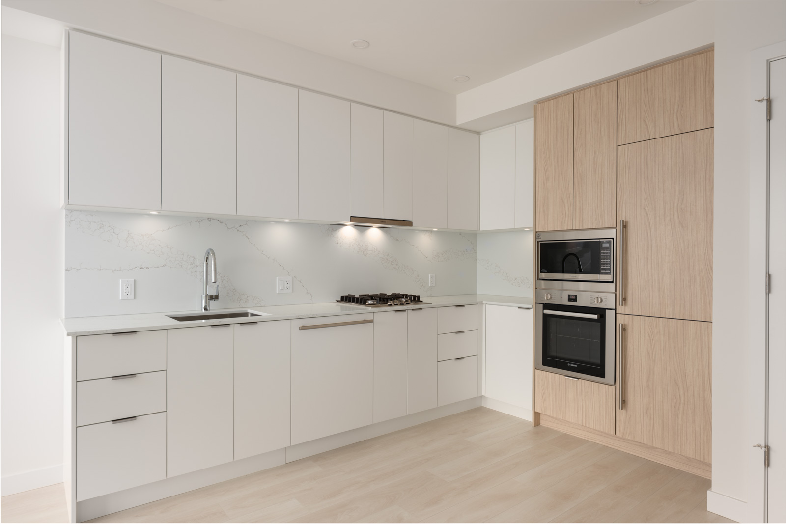 Modern kitchen with white cabinets, marble backsplash, built-in oven and microwave, gas cooktop, and light wood accents. The space is clean and minimalistic.