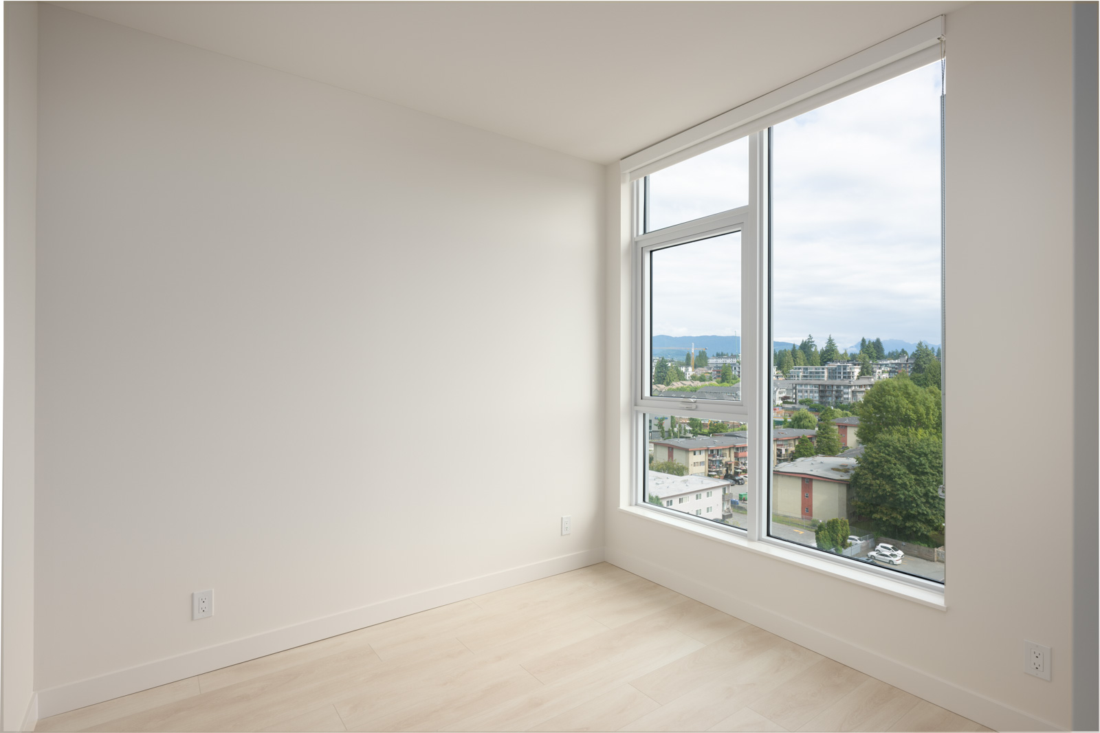 Empty room with light-colored flooring, white walls, and a large window showing an urban and tree-filled exterior view under a cloudy sky.