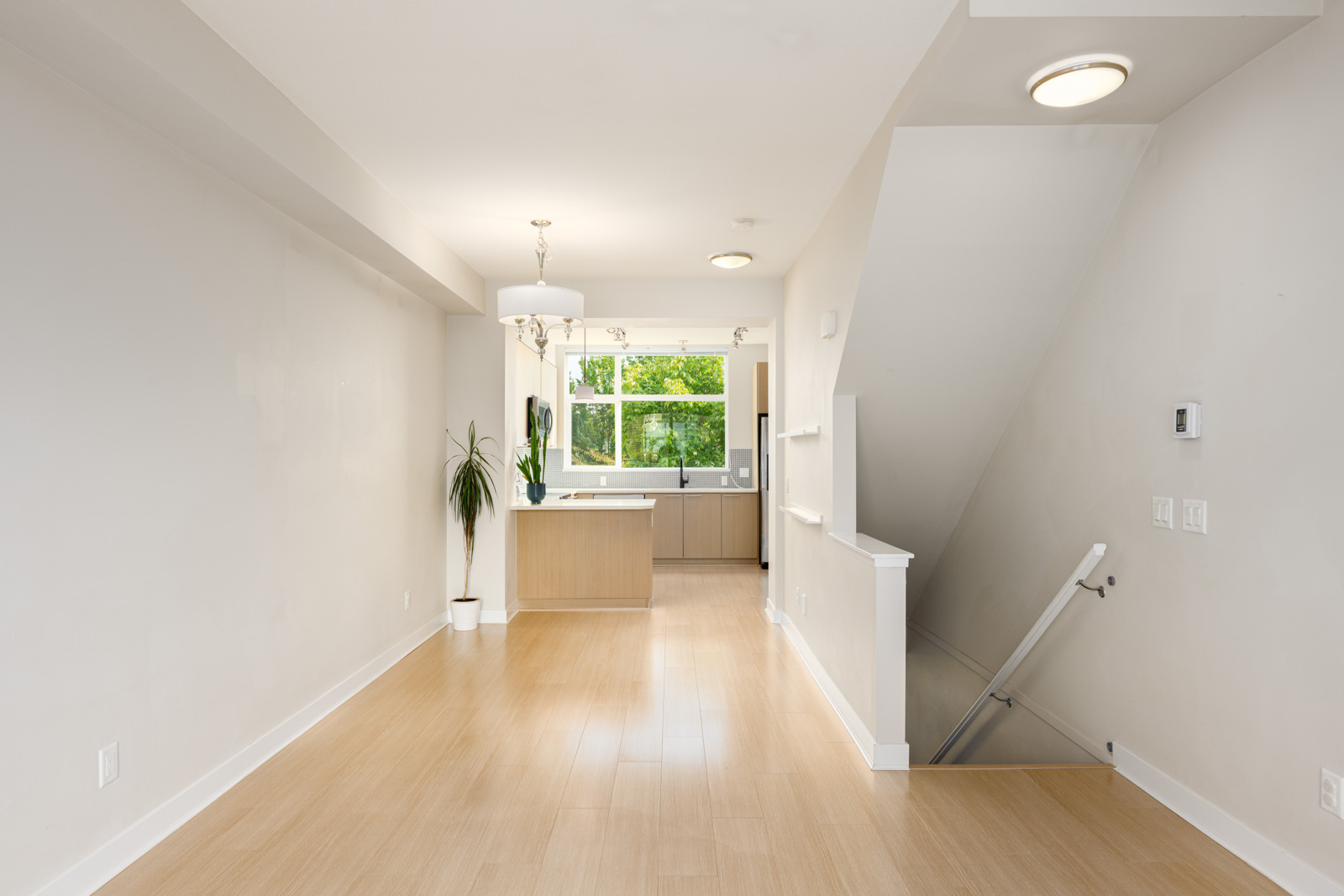 Unfurnished modern kitchen and dining area with light wood floors, white walls, a large window, a potted plant, and a stairway leading downstairs.