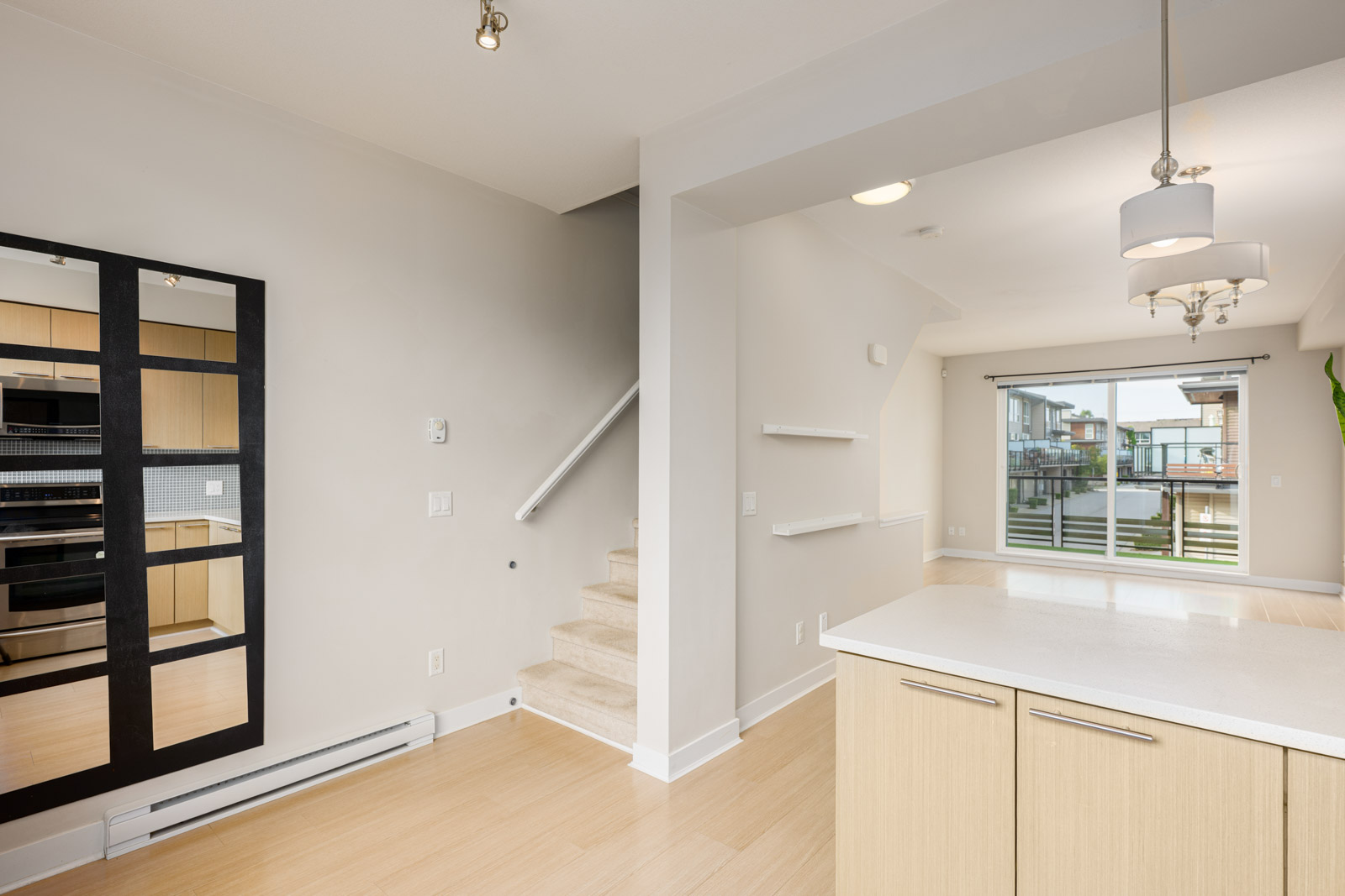 Modern open-concept kitchen and living area with light wood flooring, island, wall mirror, staircase, and large sliding glass doors leading to a balcony.